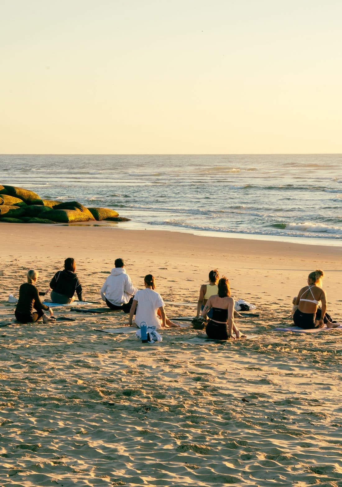 A sunrise yoga class seated on their mats on the sand, facing the sea.