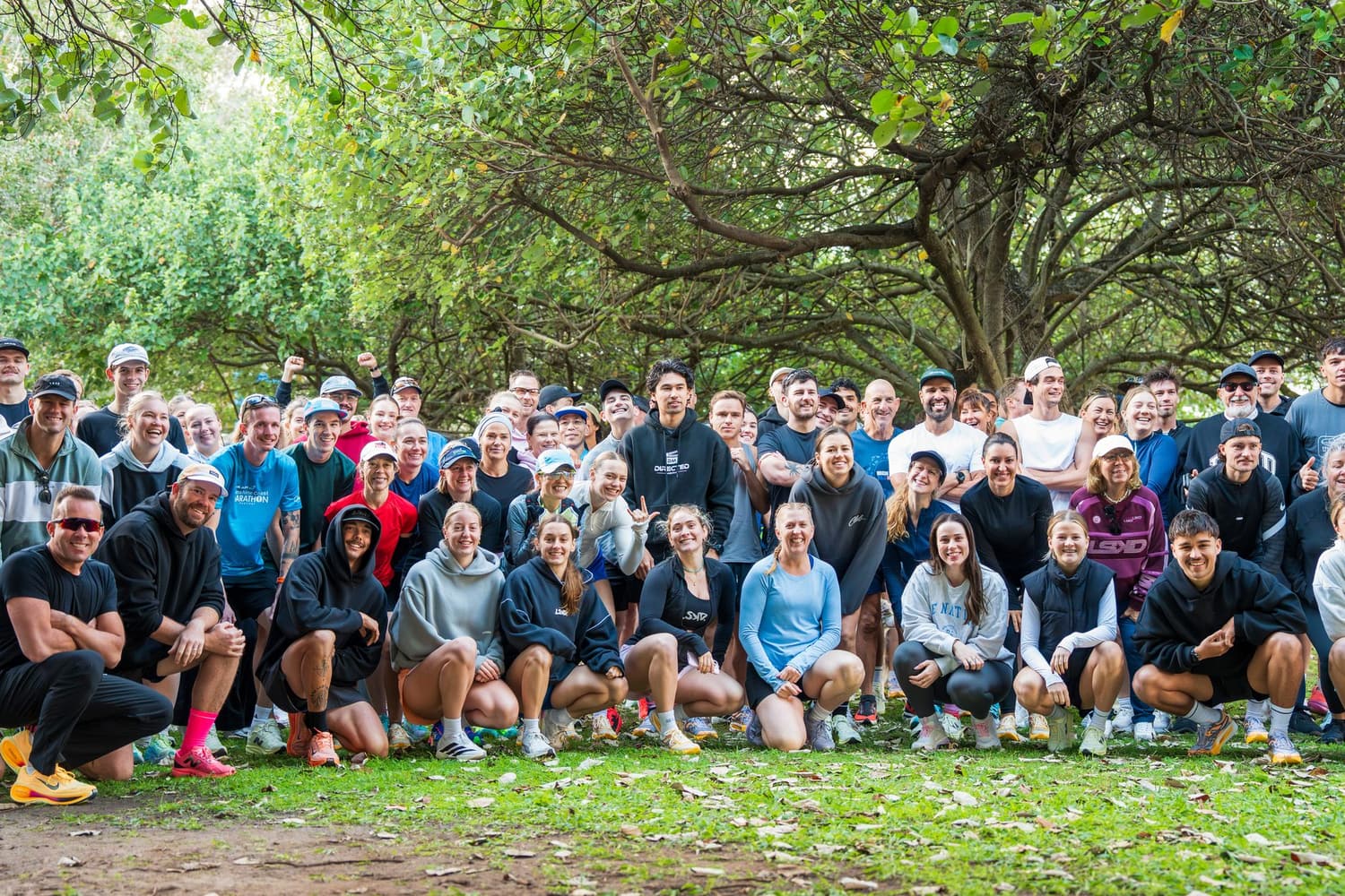 A crowd of runners gathered on the grass under the trees before a shakeout run.